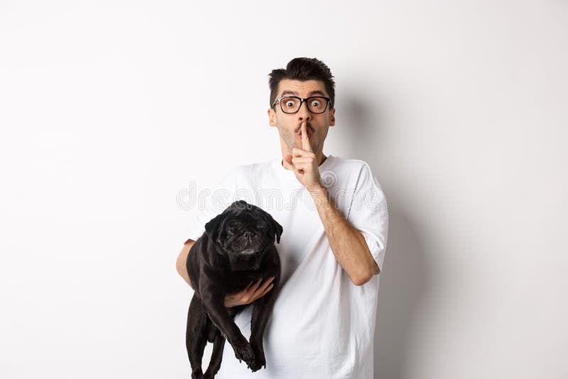 Image of funny young man holding cute black pug dog and shushing at camera, showing hush sign and staring at camera, keeping a secret, standing over white background. Image of funny young man holding cute black pug dog and shushing at camera, showing hush sign and staring at camera, keeping a secret, standing over white background.