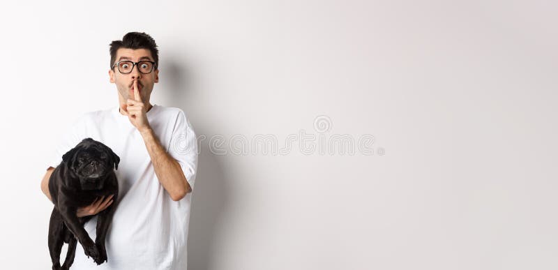 Image of funny young man holding cute black pug dog and shushing at camera, showing hush sign and staring at camera, keeping a secret, standing over white background. Image of funny young man holding cute black pug dog and shushing at camera, showing hush sign and staring at camera, keeping a secret, standing over white background.