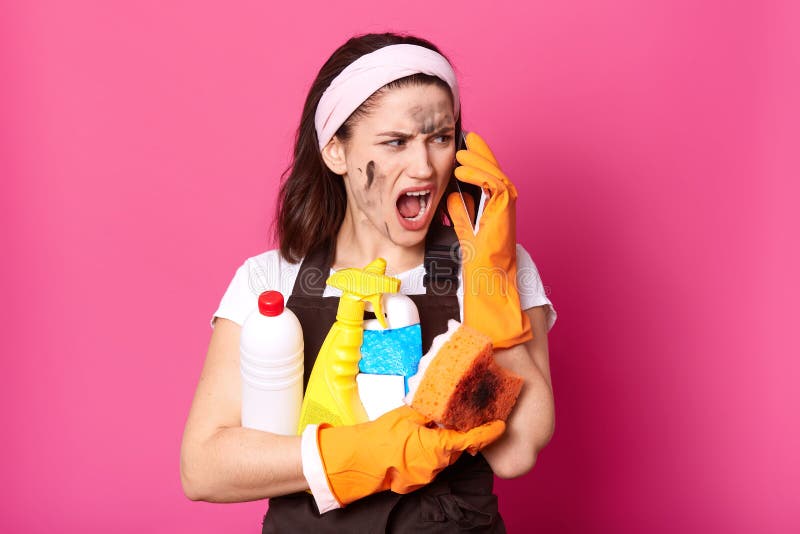 Beeld van het gillen vrouw het spreken op mobiele telefoon terwijl thuis het schoonmaken, geklede t-shirt, schort en hairband, do royalty-vrije stock afbeelding Beeld van het gillen vrouw het spreken op mobiele telefoon terwijl thuis het schoonmaken, geklede t-shirt, schort en hairband, do royalty-vrije stock afbeelding