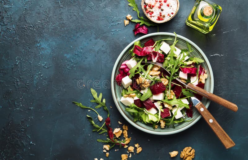 Beet or beetroot salad with fresh arugula, radicchio, soft cheese and walnuts on plate with fork, dressing and spices on blue