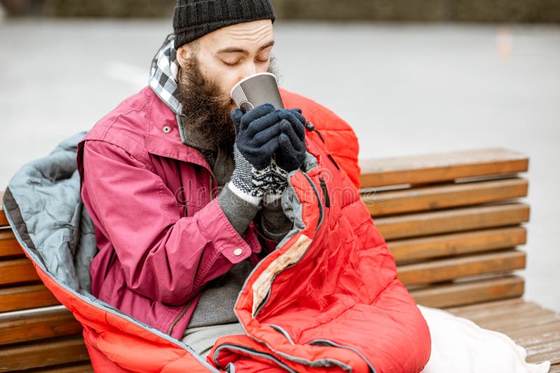 Beggar with hot drink outdoors. Homeless beggar enjoying hot drink sitting on the bench wrappped with sleeping bag outdoors royalty free stock photo Beggar with hot drink outdoors. Homeless beggar enjoying hot drink sitting on the bench wrappped with sleeping bag outdoors royalty free stock photo