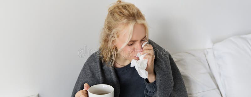 Concept of flu and quarantine. Close up portrait of young woman feeling sick, caught a cold, sneezing and blowing nose in napkin, holding warm tea in hand. Concept of flu and quarantine. Close up portrait of young woman feeling sick, caught a cold, sneezing and blowing nose in napkin, holding warm tea in hand.