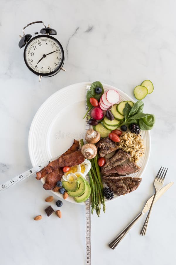 16:8 Intermittent fasting weigh loss concept. Various low carb foods on a white plate with a portion empty and clock to illustrate a condensed eating window. 16:8 Intermittent fasting weigh loss concept. Various low carb foods on a white plate with a portion empty and clock to illustrate a condensed eating window.