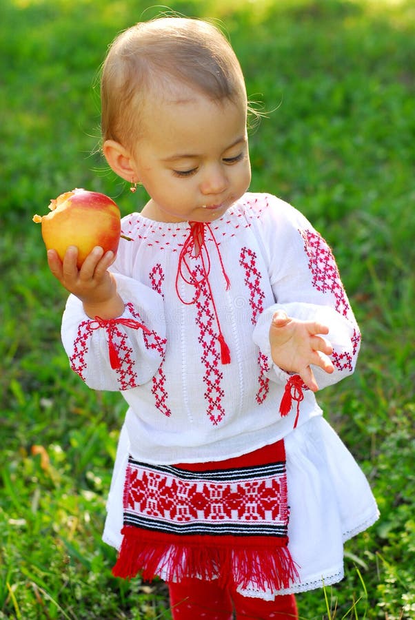 Behandla som ett barn den iklädda traditionella dräkten för flickan och att äta ett äpple arkivfoto Behandla som ett barn den iklädda traditionella dräkten för flickan och att äta ett äpple arkivfoto