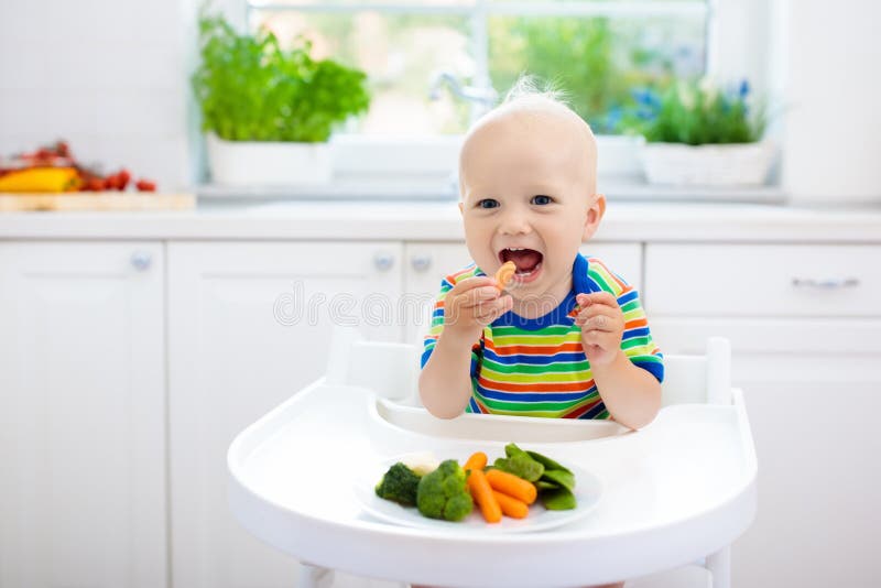 Cute baby eating vegetables in white kitchen. Infant weaning. Little boy trying solid food, organic broccoli, cauliflower, carrot and green peas. Healthy nutrition for kids. Child biting carrot. Cute baby eating vegetables in white kitchen. Infant weaning. Little boy trying solid food, organic broccoli, cauliflower, carrot and green peas. Healthy nutrition for kids. Child biting carrot.