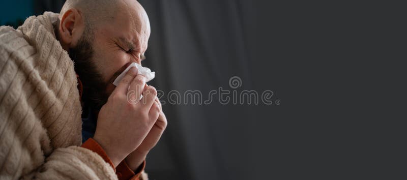 Needs treatment and medicines. Sneezing, runny nose, cough. Ill man sits on the couch at home, sneezing in a napkin. Banner. Copy space. Needs treatment and medicines. Sneezing, runny nose, cough. Ill man sits on the couch at home, sneezing in a napkin. Banner. Copy space
