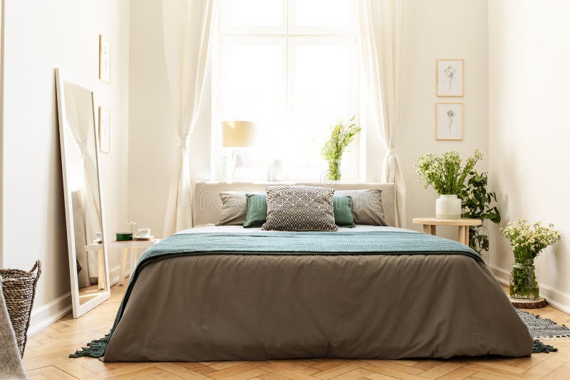 Beige, groen en grijs slaapkamerbinnenland in een huurkazerne met een bed tegen een zonnig venster en bossen van wilde bloemen Ec stock foto Beige, groen en grijs slaapkamerbinnenland in een huurkazerne met een bed tegen een zonnig venster en bossen van wilde bloemen Ec stock foto