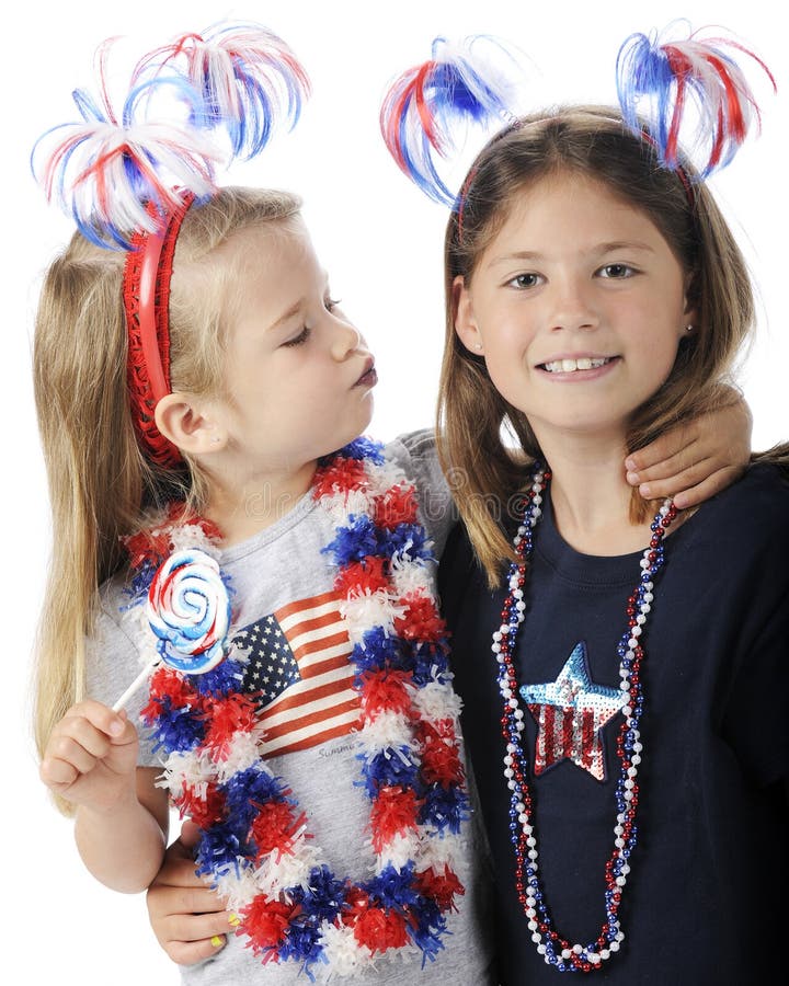 An adorable preschooler giving her sister a kiss with lips turned blue from her lollipop. Both girls wear American patriot accessories. On a white background. An adorable preschooler giving her sister a kiss with lips turned blue from her lollipop. Both girls wear American patriot accessories. On a white background.