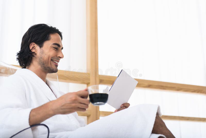 Joyful young man is relaxing in morning. He is drinking coffee and reading book. Man is sitting in bathrobe and smiling. Joyful young man is relaxing in morning. He is drinking coffee and reading book. Man is sitting in bathrobe and smiling