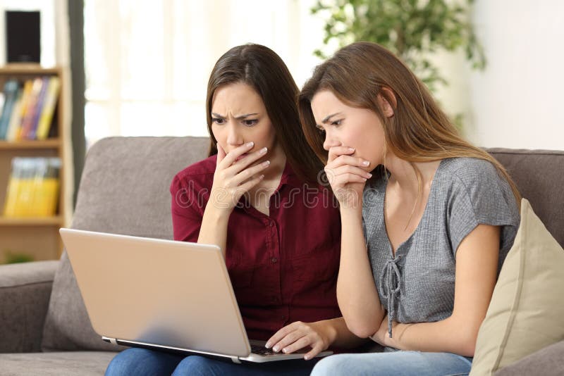 Two worried friends watching media content on line with a pc sitting on a sofa in the living room in a house indoor. Two worried friends watching media content on line with a pc sitting on a sofa in the living room in a house indoor