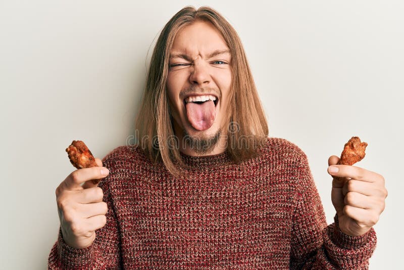 Bel homme caucasien avec de longs cheveux mangeant des ailes de poulet en sortant la langue heureux avec l'expression drôle images libres de droits