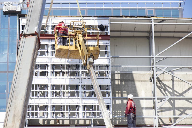 Construction workers in crane basket installing cladding metal structure on a building facade wall