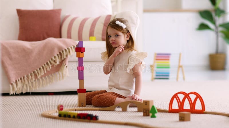 Bella bambina di rossa che gioca sul tappeto a casa impilando dei colorati blocchi di legno che costruiscono una torre archivi video