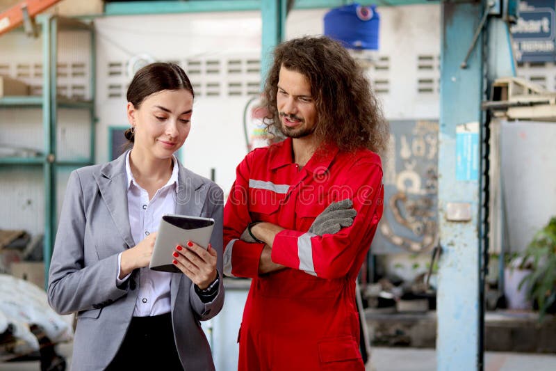 Bella clienta mujer sostiene tableta digital discutiendo con mecánico de uniforme rojo trabajador automotor inspeccionando el auto fotografía de archivo
