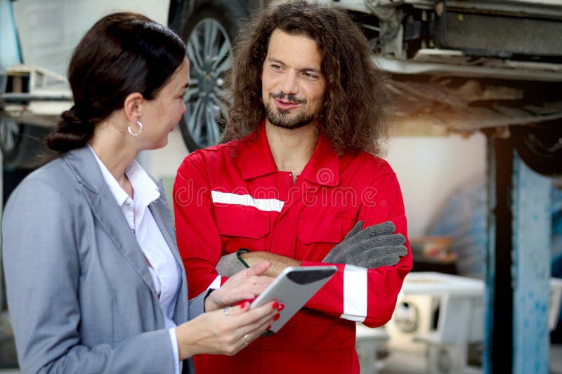 Bella clienta mujer sostiene tableta digital discutiendo con mecánico de uniforme rojo trabajador automotor inspeccionando el auto imagenes de archivo