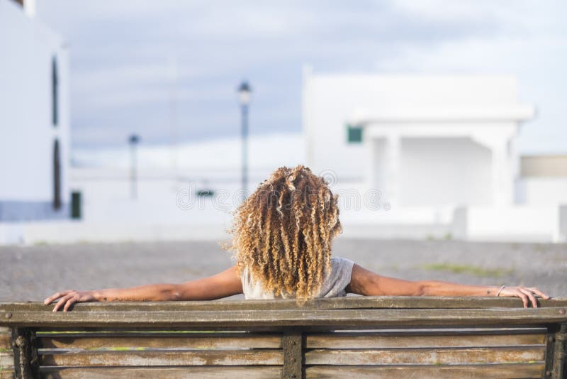 Bella donna abbronzata dei capelli ricci pelle nera osservata dalla parte posteriore che si siede su un banco