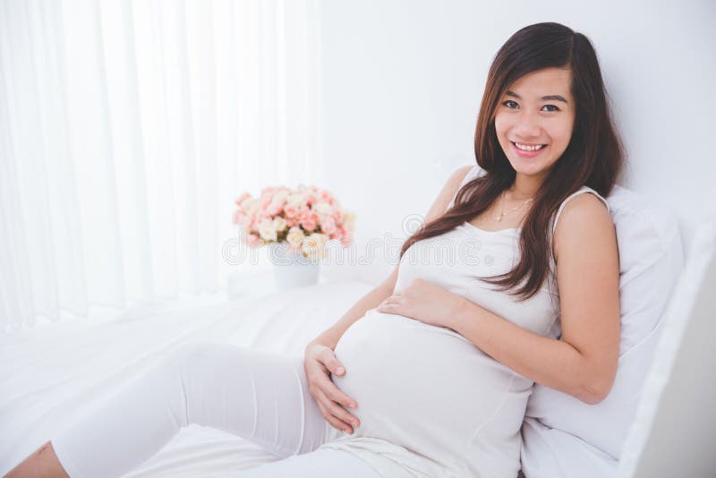 Bella donna asiatica incinta che si appoggia un letto, sorridente fotografie stock libere da diritti Bella donna asiatica incinta che si appoggia un letto, sorridente fotografie stock libere da diritti