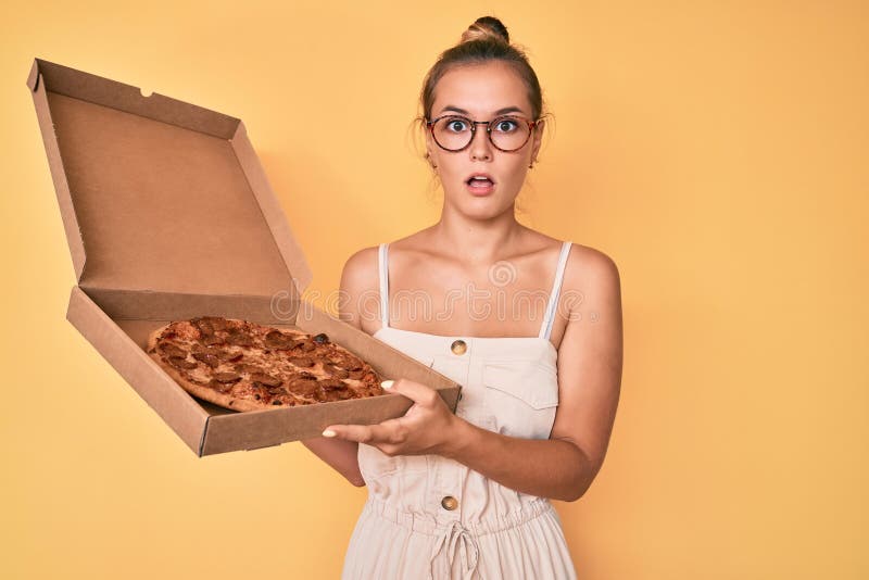 Beautiful caucasian woman holding tasty pepperoni pizza in shock face, looking skeptical and sarcastic, surprised with open mouth. Beautiful caucasian woman holding tasty pepperoni pizza in shock face, looking skeptical and sarcastic, surprised with open mouth