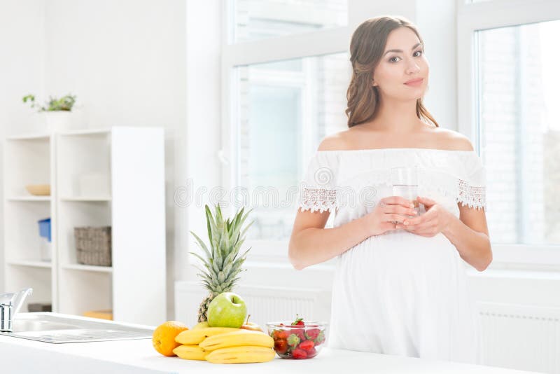 Bella donna incinta nella cucina che ha un bicchiere d'acqua immagini stock libere da diritti Bella donna incinta nella cucina che ha un bicchiere d'acqua immagini stock libere da diritti
