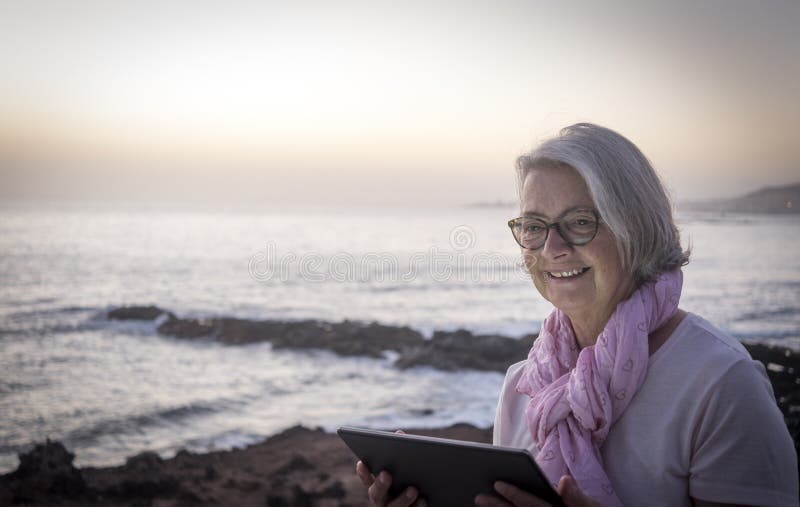 Bella e sorridente donna di capelli grigi seduta vicino alla spiaggia a navigare sulla rete con una tavoletta. oscuramento e orizz immagini stock