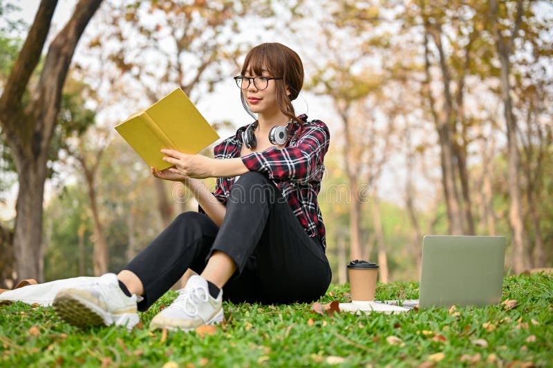 Beautiful and calm young Asian female college student in casual clothes sits on the grass and reads a book,spending her free time on the weekend at the park. Beautiful and calm young Asian female college student in casual clothes sits on the grass and reads a book,spending her free time on the weekend at the park