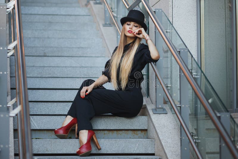 Bella giovane donna alla moda che posa nella serie nera, scarpe con i tacchi alti e black hat rossi Stile di moda Cenni storici u fotografia stock libera da diritti Bella giovane donna alla moda che posa nella serie nera, scarpe con i tacchi alti e black hat rossi Stile di moda Cenni storici u fotografia stock libera da diritti