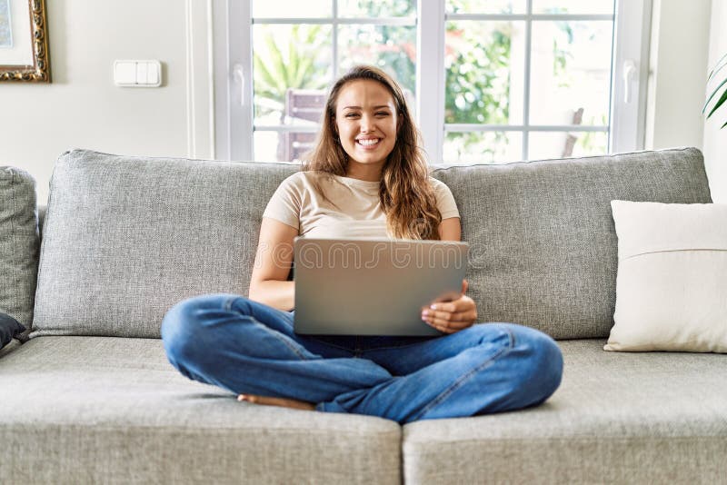 Bella giovane donna brunetta seduta sul divano che usa il computer portatile a casa con un sorriso allegro e fresco sulla faccia fotografie stock