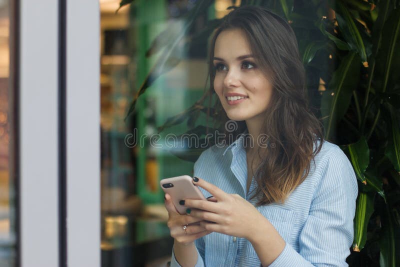 Bella giovane donna caucasica sveglia nel caffè, facendo uso del telefono cellulare e della condizione vicino a sorridere della f fotografia stock Bella giovane donna caucasica sveglia nel caffè, facendo uso del telefono cellulare e della condizione vicino a sorridere della f fotografia stock