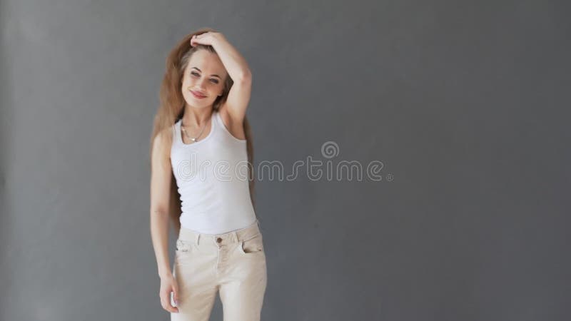 Bella giovane donna con capelli biondi sani In una canottiera sportiva bianca su un fondo grigio, posante sulla macchina fotograf