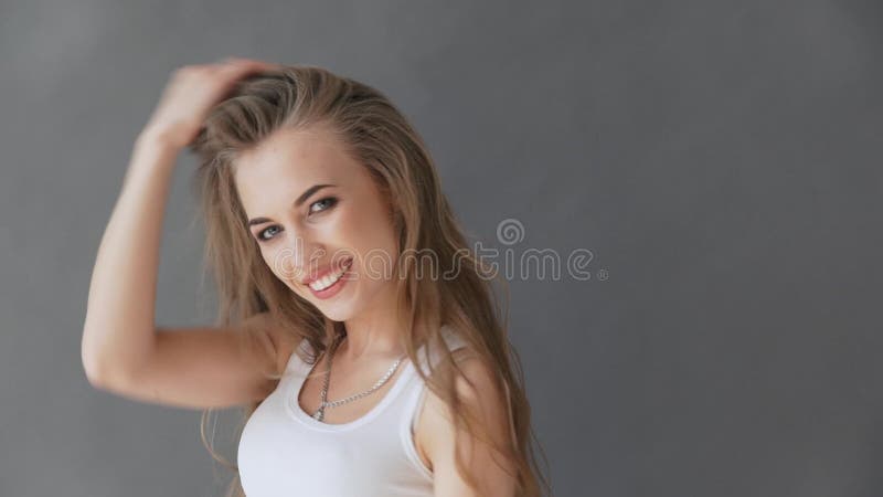 Bella giovane donna con capelli biondi sani In una canottiera sportiva bianca su un fondo grigio, posante sulla macchina fotograf
