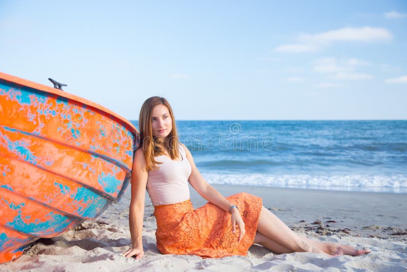 Bella giovane donna dai capelli rossi in gonna che si rilassa sulla spiaggia vicino alla barca arancio fotografia stock Bella giovane donna dai capelli rossi in gonna che si rilassa sulla spiaggia vicino alla barca arancio fotografia stock
