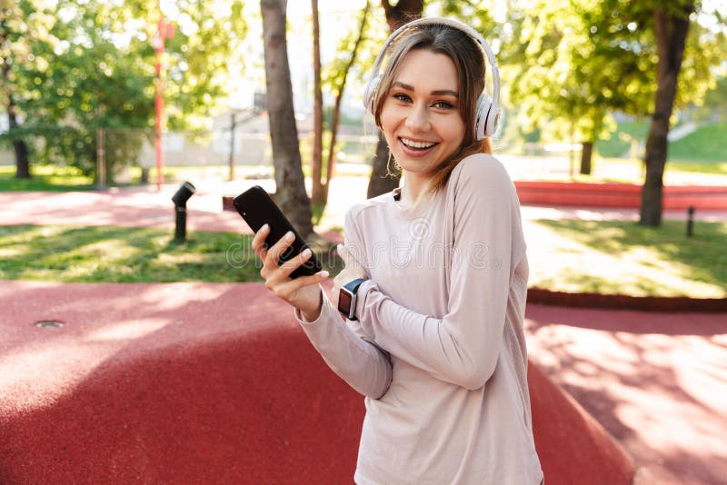 Bella giovane donna sportiva di fitness che posava all'aperto in un parco ascoltando musica con cuffie con cellulare fotografia stock libera da diritti Bella giovane donna sportiva di fitness che posava all'aperto in un parco ascoltando musica con cuffie con cellulare fotografia stock libera da diritti