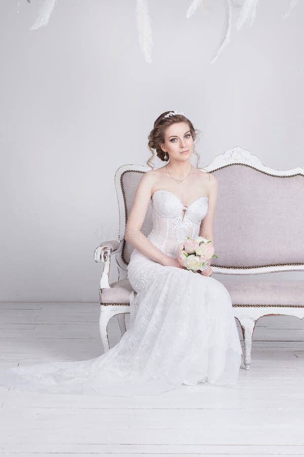 Bella giovane sposa in un vestito da sposa lussuoso dal pizzo Si siede su un sofà d'annata bianco fotografie stock Bella giovane sposa in un vestito da sposa lussuoso dal pizzo Si siede su un sofà d'annata bianco fotografie stock