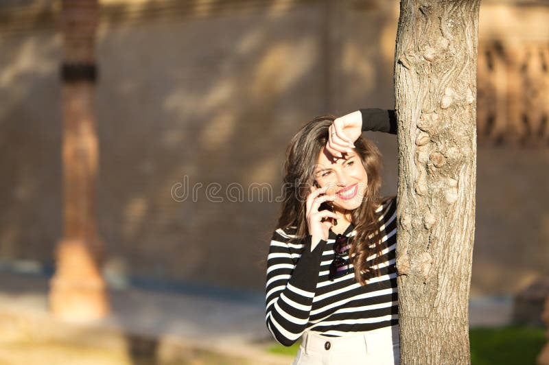 Bella joven de cabello largo y marrón con una camiseta de rayas blancas y negras inclinada contra un árbol que habla en su móvil fotos de archivo