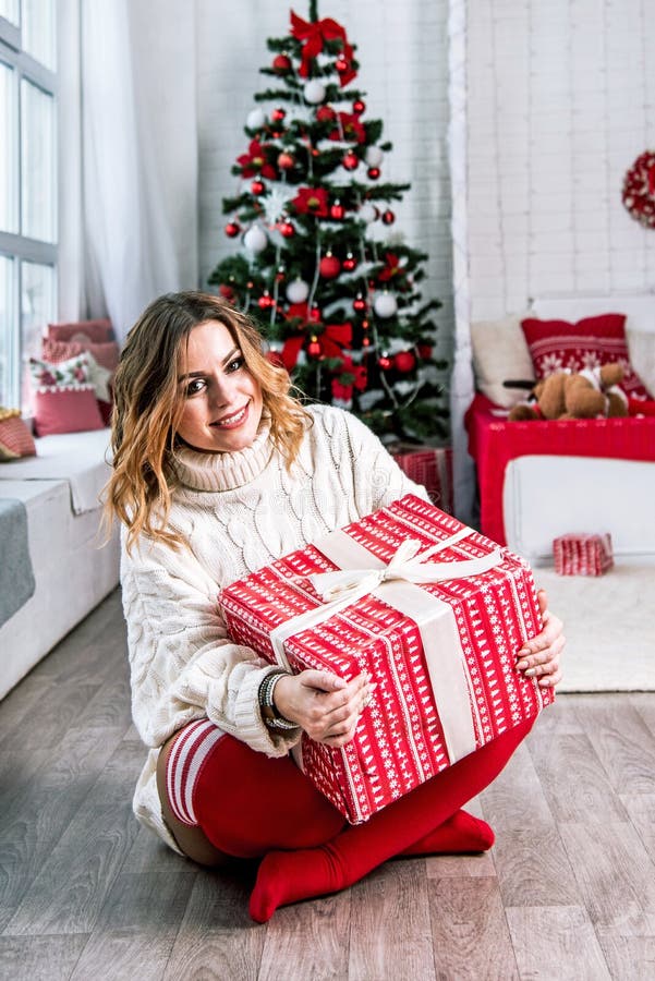 Bella madre alegre con suéter de punto blanco y piernas rojas con una gran caja de regalo en un arco contra un fondo de árbol de n foto de archivo