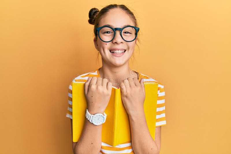 Bella morena pequeña niña con gafas y sosteniendo un libro sonriendo y riéndose fuerte porque chiste loco gracioso imágenes de archivo libres de regalías