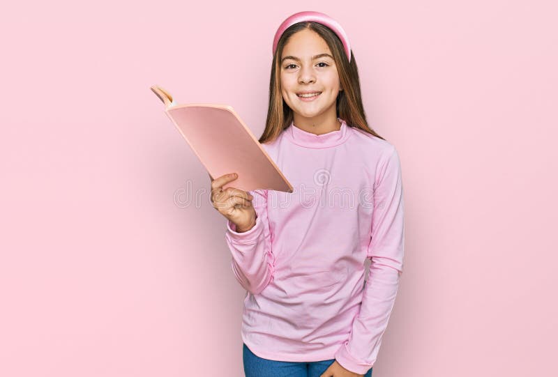 Bella morena pequeña niña leyendo un libro con aspecto positivo y feliz de pie y sonriendo con una sonrisa segura mostrando diente imagenes de archivo