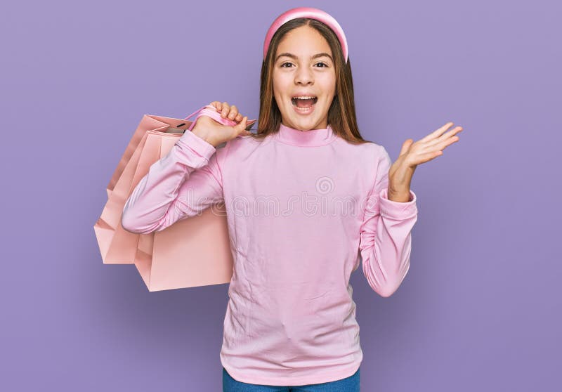 Bella morena pequeña niña sujeta bolsas de compras celebrando la victoria con sonrisa feliz y expresión del ganador con las manos foto de archivo libre de regalías
