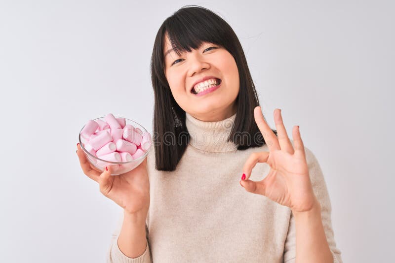 Bella mujer china sosteniendo un tazón con malvaviscos sobre fondo blanco aislado haciendo señas bien con los dedos excelente símb imagen de archivo