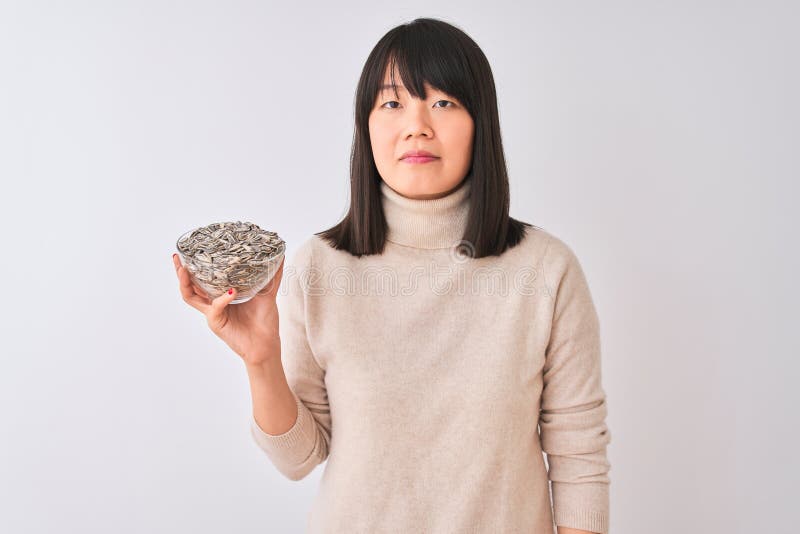 Bella mujer china sosteniendo un tazón con semillas de girasol sobre fondo blanco aislado con una expresión segura en inteligente fotos de archivo libres de regalías