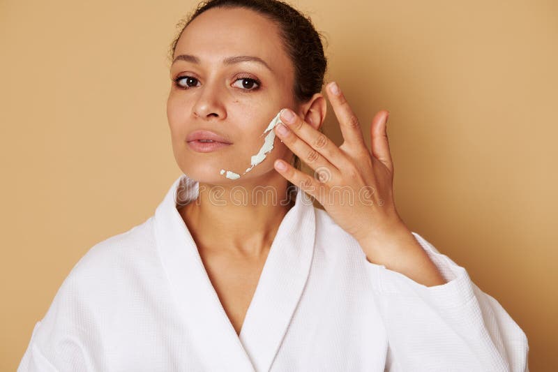 Bella mujer de raza mixta aplicando una máscara de belleza en su cara. retrato de la imagen en el fondo beige con espacio de copia imagen de archivo libre de regalías