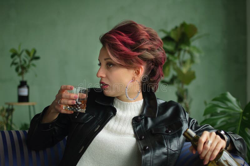 Bella Mujer Sexy Con Pelo Rosa Oscuro En De Cuero Negro Está Bebiendo Whisky De Un Vaso Sentado En Un Sofá Azul En Foto de archivo - Imagen de persona, presionado: