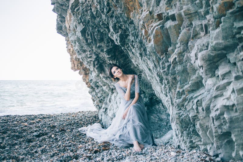 Bella ragazza castana in vestito blu che si siede vicino alle rocce, mare, sorridente, fotografia stock libera da diritti Bella ragazza castana in vestito blu che si siede vicino alle rocce, mare, sorridente, fotografia stock libera da diritti