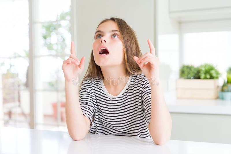 Bella ragazza che indossa una camicia a strisce stupita e sorpresa guardando in alto e puntando con le dita e alzando le braccia immagine stock