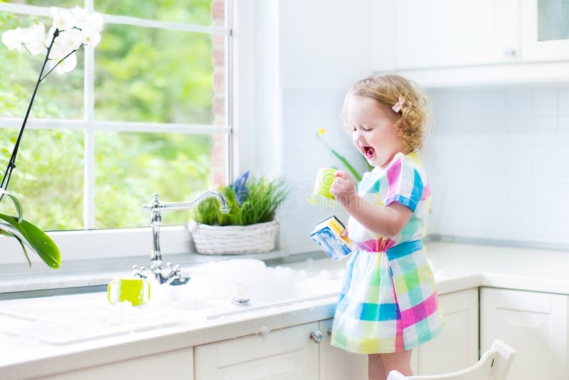 Cute curly toddler girl in a colorful dress washing dishes, cleaning with a sponge and playing with foam in the sink in a beautiful sunny white kitchen with a garden view window in a modern home. Cute curly toddler girl in a colorful dress washing dishes, cleaning with a sponge and playing with foam in the sink in a beautiful sunny white kitchen with a garden view window in a modern home