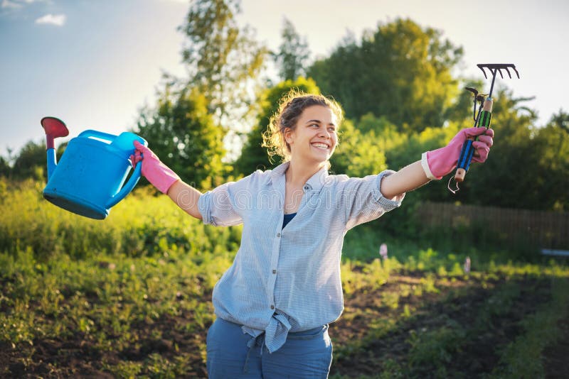 Beautiful young woman girl with a watering can and a rake in her hands against the background of the garden, work in the garden and eco-friendly lifestyle. Beautiful young woman girl with a watering can and a rake in her hands against the background of the garden, work in the garden and eco-friendly lifestyle