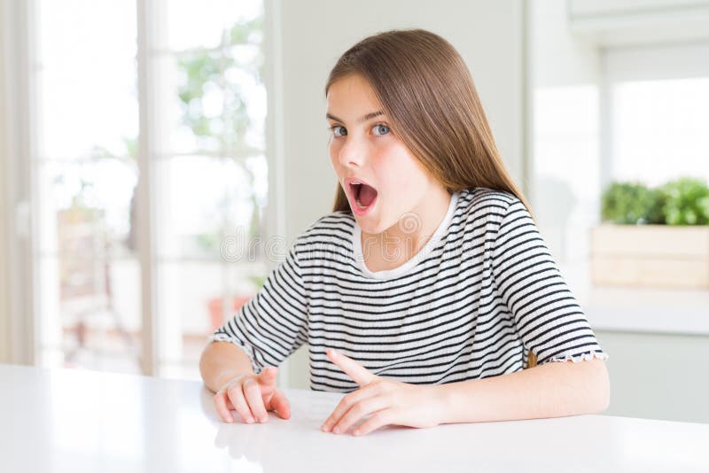 Bella ragazzina con una camicia a strisce spaventata e scioccata dall'espressione a sorpresa paura e faccia eccitata immagine stock