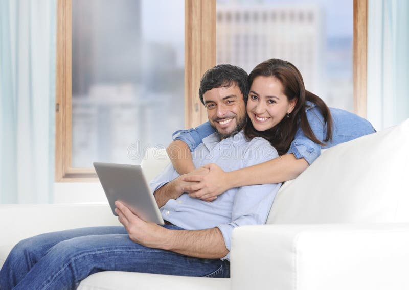 Belle coppie latine nell'amore che si trova insieme sullo strato del sofà del salone che gode per mezzo della compressa digitale fotografia stock Belle coppie latine nell'amore che si trova insieme sullo strato del sofà del salone che gode per mezzo della compressa digitale fotografia stock