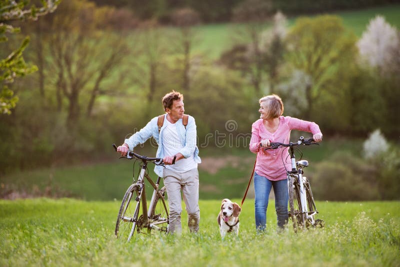 Belle coppie senior con le biciclette ed il cane fuori nella natura di primavera fotografie stock Belle coppie senior con le biciclette ed il cane fuori nella natura di primavera fotografie stock