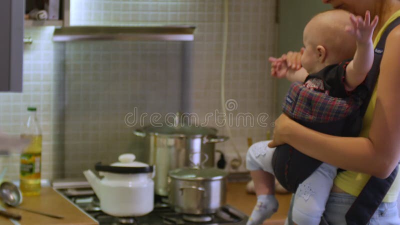 Belle femme avec un bébé garçon assis dans une soupe de cuisson porte-bébé pour un dîner à la cuisine. foyer mère